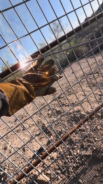 A gloved hand welding wire mesh onto a steel pipe frame, sparks flying, desert dirt and blue sky behind.