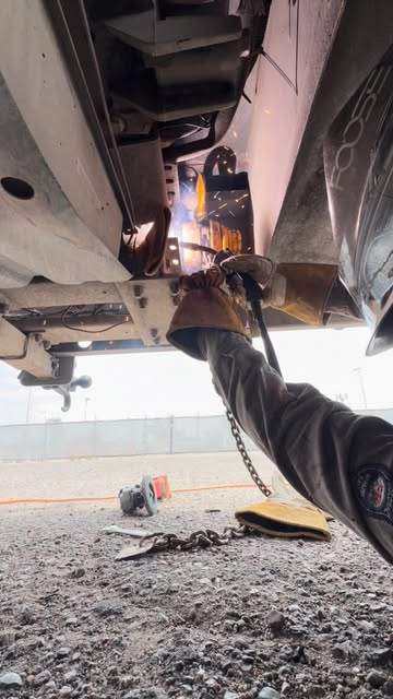 A welder lying under a pickup in a gravel lot, welding the frame with sparks flying.