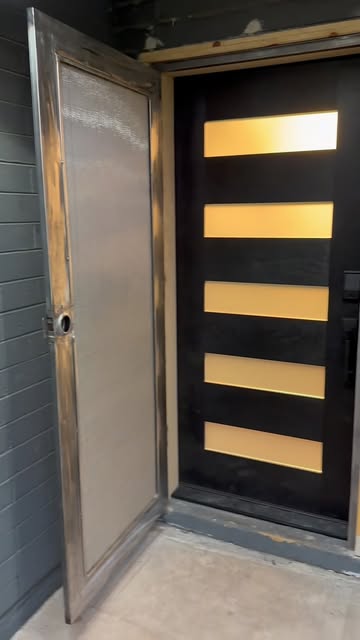 A custom steel security door with a full screen insert, swung open in front of a modern black front door with glowing glass lites.
