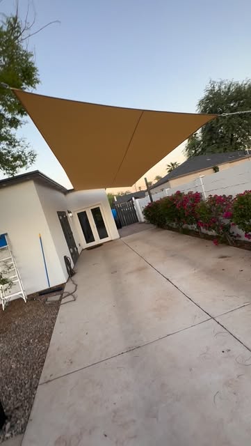 A tan shade sail stretched over a concrete driveway beside a white casita and bougainvillea at dusk.