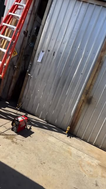 A corrugated steel sliding shop door in the sun, a red welder on the concrete and an orange ladder beside it.