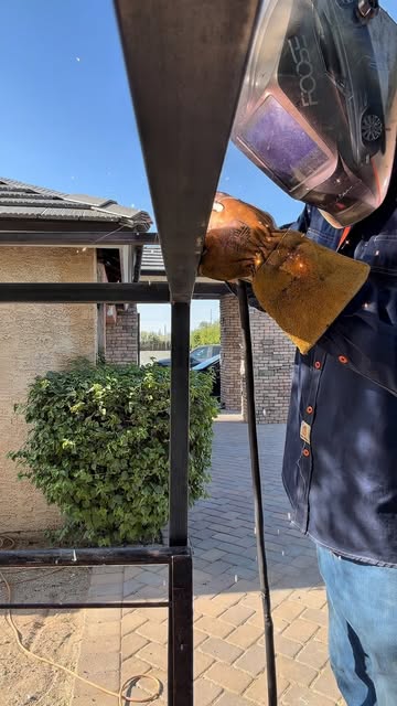 A welder in a helmet and leather gloves welding a square tube rack in a paver driveway beside a stucco house.